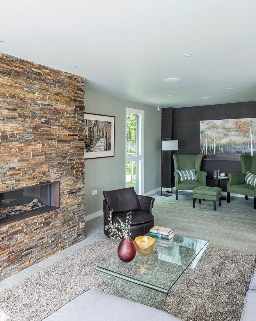 Glass table on pale grey rug, fireplace integrated into stone wall and green armchairs in seating area in open-plan interior