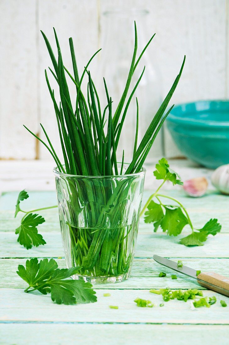 Schnittlauch im Wasserglas, daneben Petersilie und Knoblauch auf Holzuntergrund