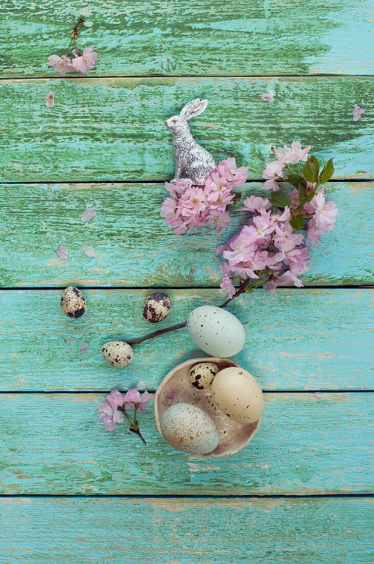 Easter eggs and quail's eggs in bowl, silver Easter bunny and cherry blossom on wooden surface