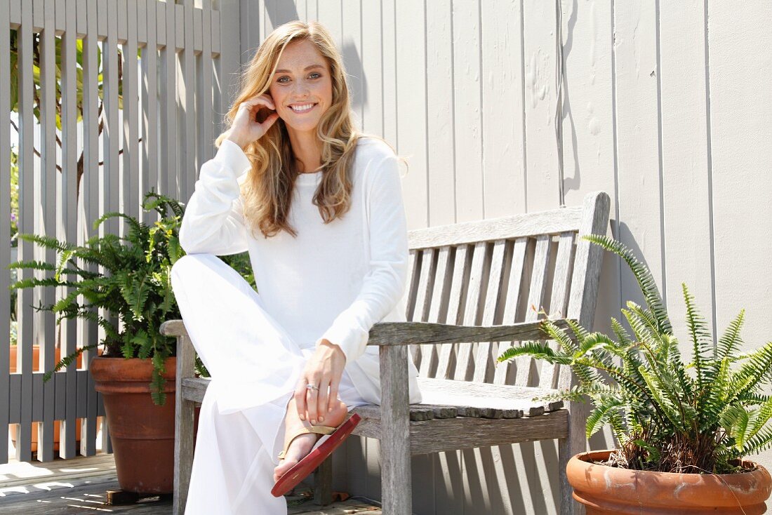 A blonde woman wearing loose-fitting, white clothing sitting on the bench on a terrace