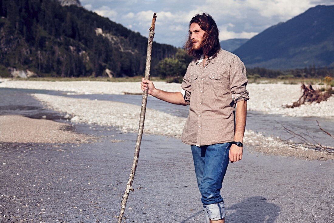 A young man with a beard and long hair walking along a gravelly river bed
