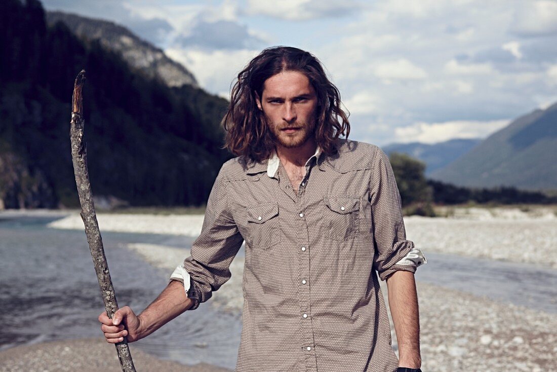 A young man with a beard and long hair walking along a gravelly river bed