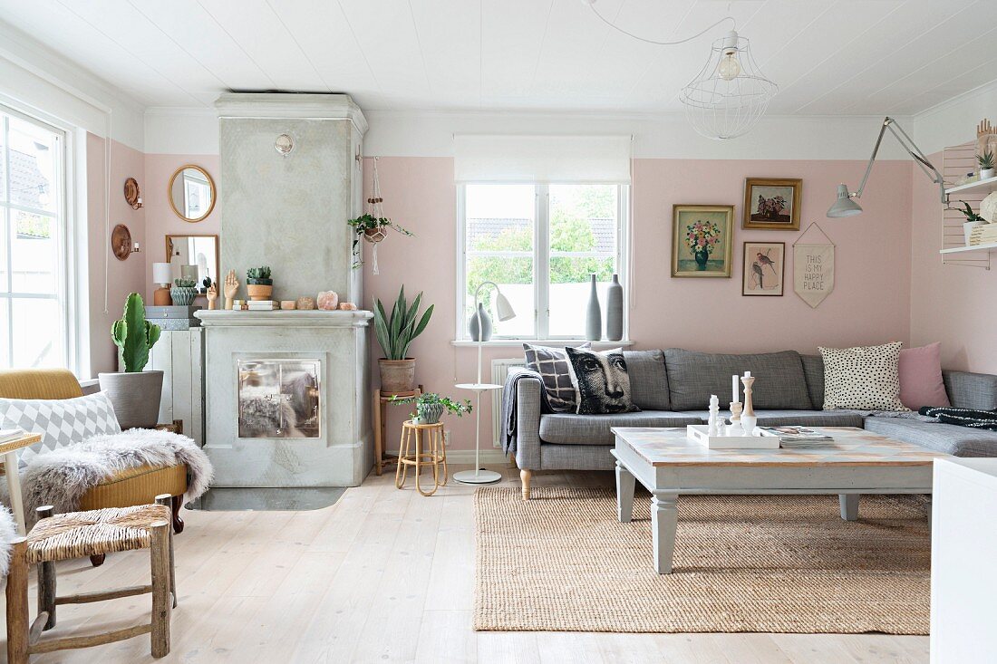 Gemütliches Wohnzimmer mit hellgrau lackierter Couchtisch und Polstersofa in Zimmerecke, seitlich Kamin aus Beton mit verchromten Türen an rosa getönter Wand