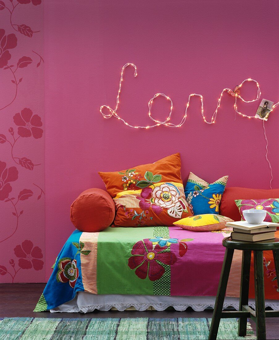 Tagesdecke und Kissen mit Blütenmotiven auf Sofa vor pinkfarbener Wand mit Lichterkette und Schriftzug Love