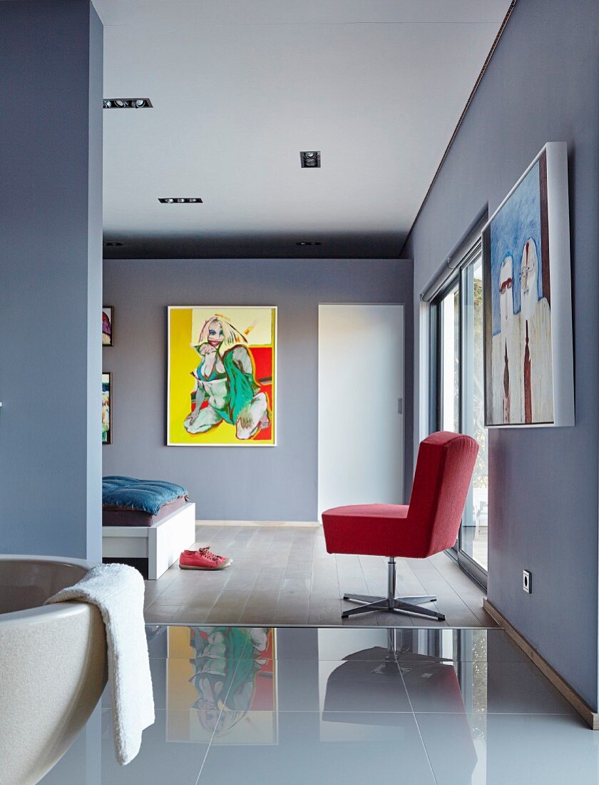 Bad Ensuite mit glänzendem Fliesenboden vor blau-grauem Schlafbereich mit rotem Drehsessel und moderner Malerei