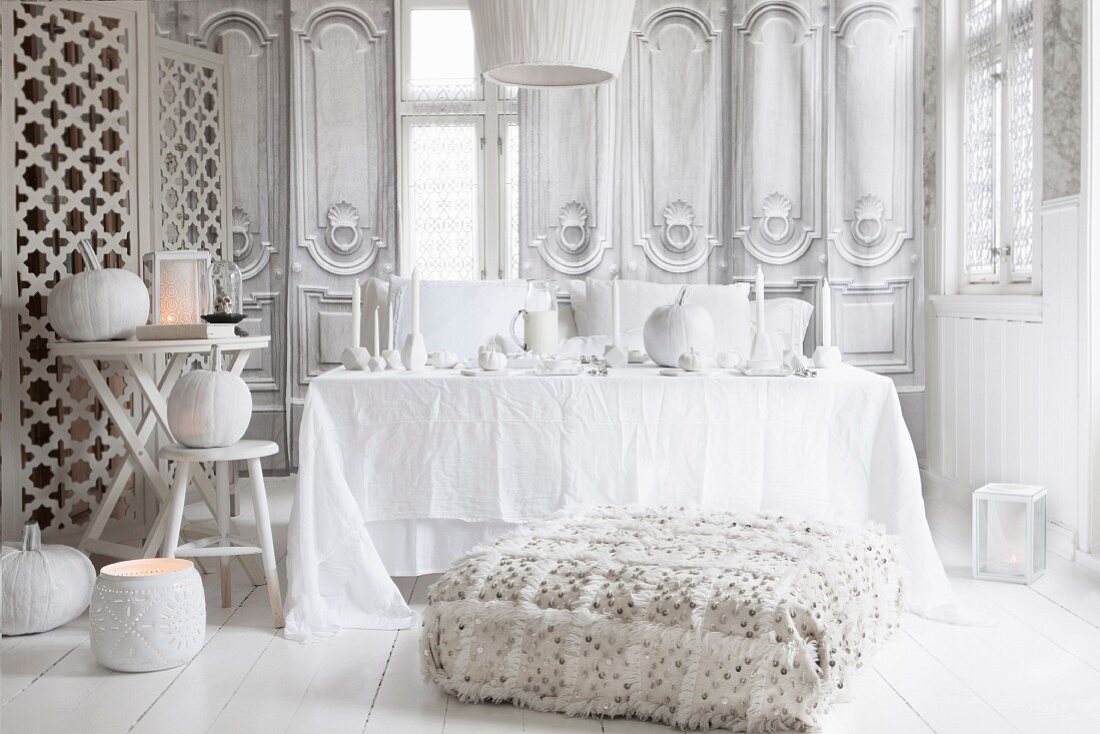 Stilvolles Halloween Arrangement in Weiß, gemütliches Sitzpolster vor dekoriertem Tisch, seitlich Dekokürbisse und Windlichter