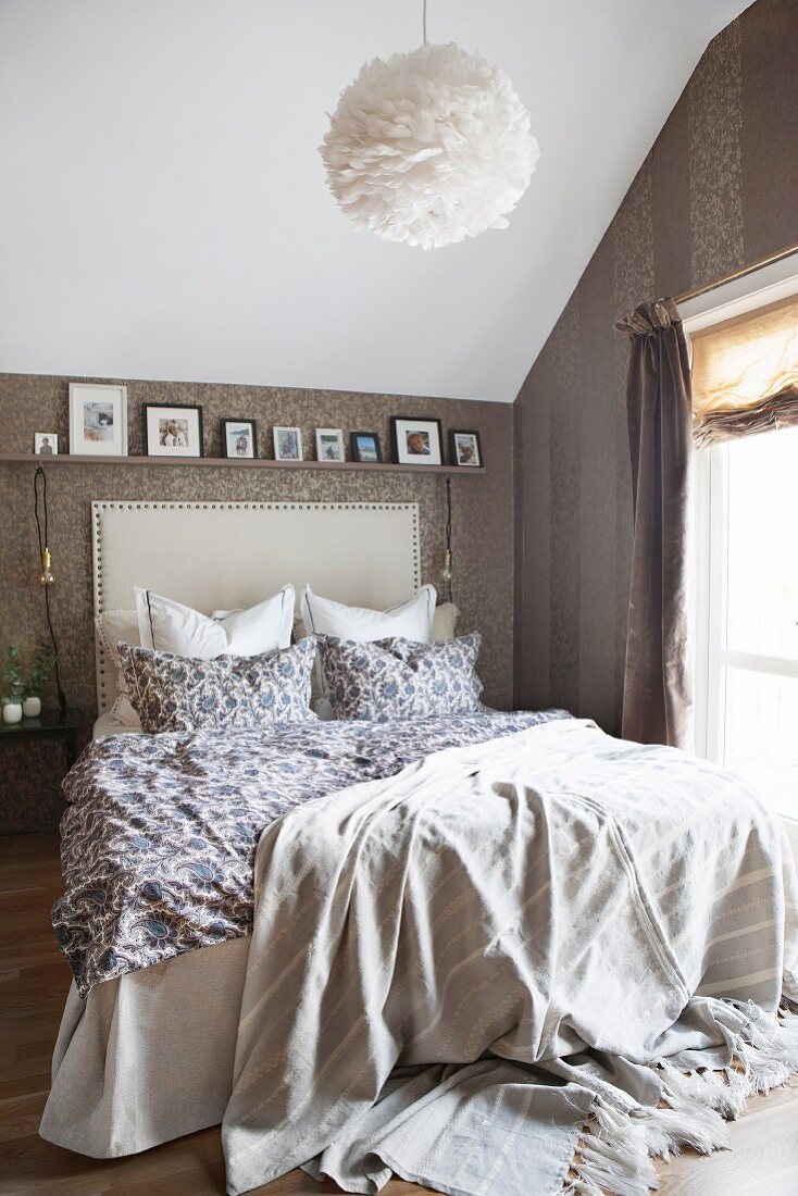 Bed with upholstered headboard under sloping ceiling in attic bedroom with patterned wallpaper and shelf of pictures