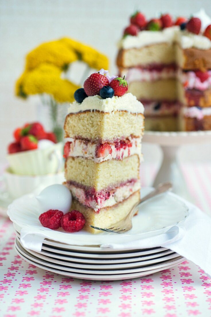 Eton-Mess-Schichtkuchen mit Erdbeeren, Himbeeren und Heidelbeeren, angeschnitten, mit Stück auf Teller