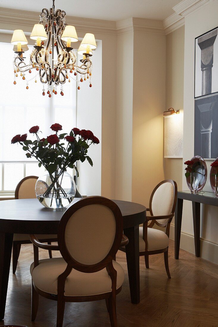 Chandelier above vase of red roses on round dining table and upholstered armchairs with oval backrests