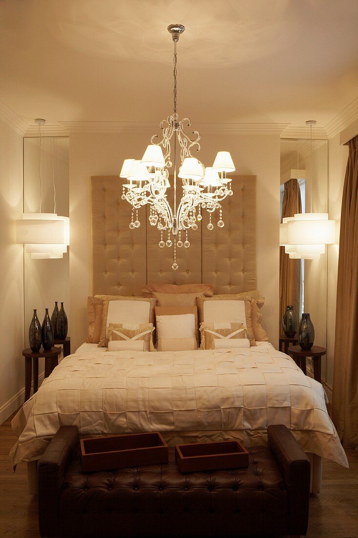 Crystal chandelier with lampshades above elegant double bed with tall button-tufted headboard flanked by vases in front of mirrors