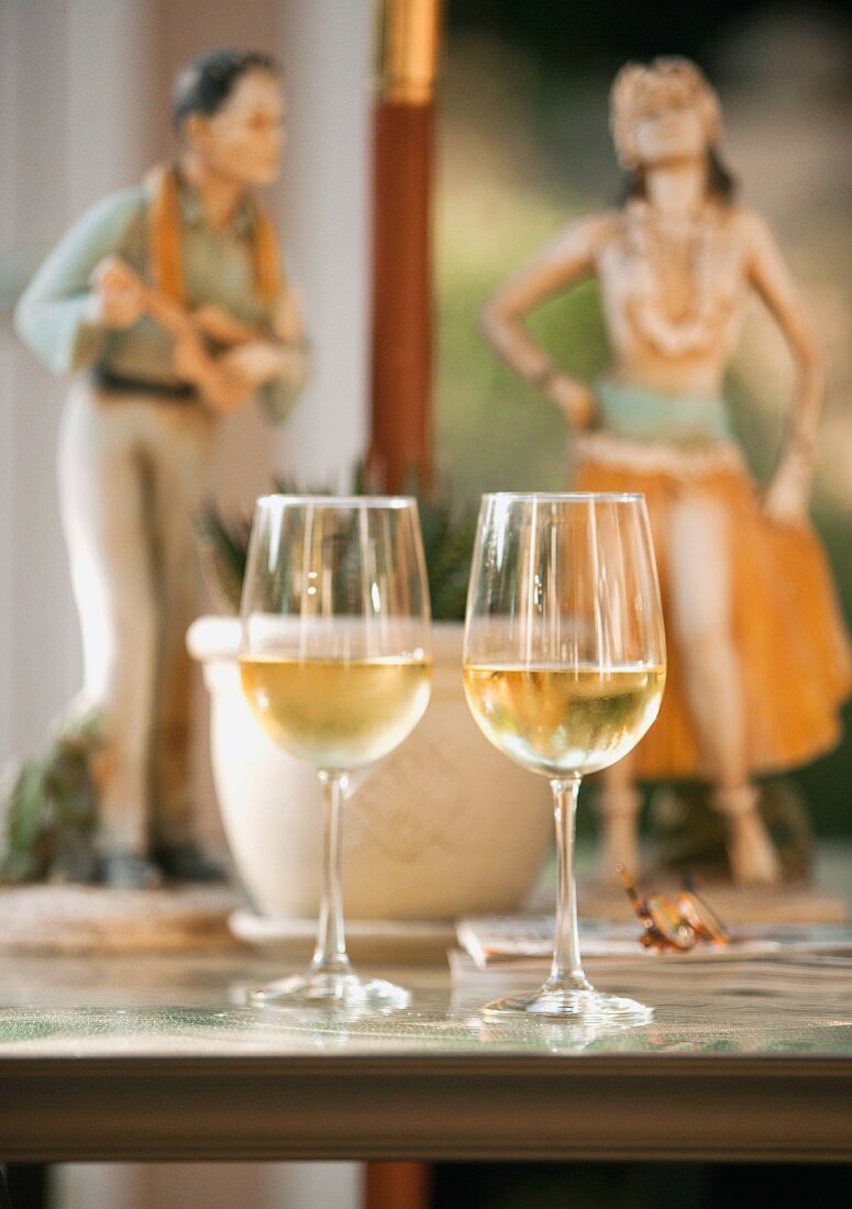 Two glasses of white wine on a table with two Hawaiian statues in the background