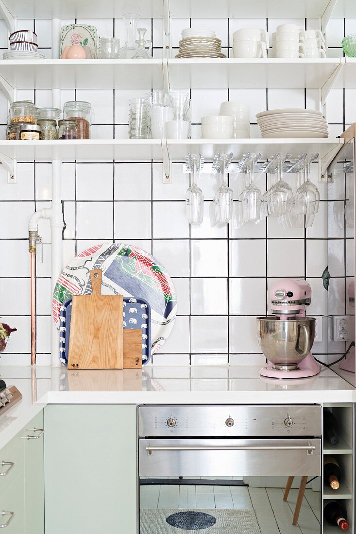 L-shaped kitchen counter with white worksurface below crockery on shelves on white-tiled wall