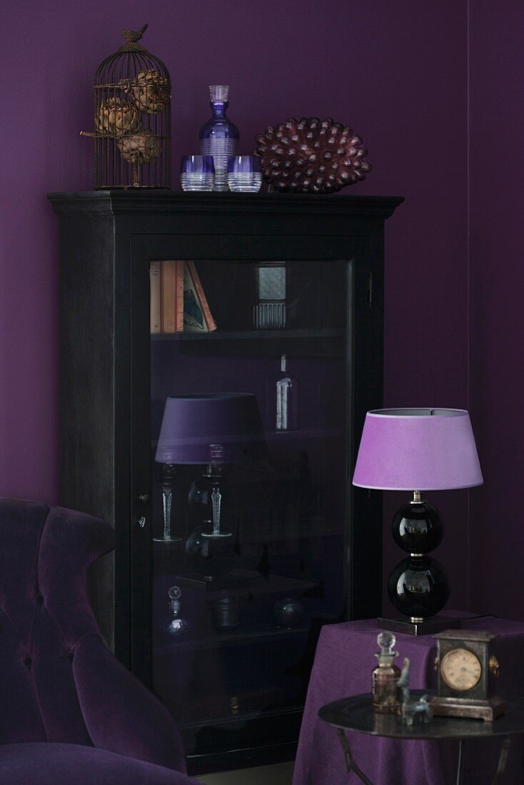 Table lamp with lilac lampshade in front of antique, glass-fronted cabinet against aubergine wall