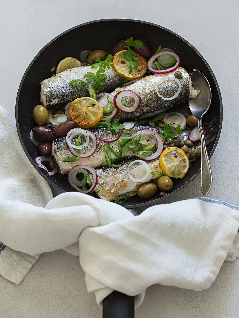 Fried fish in a pan with olives, capers, onions and lemons