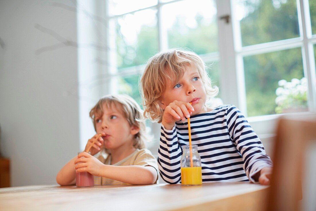 Zwei Kinder sitzen am Tisch & trinken mit Strohhalmen Smoothies