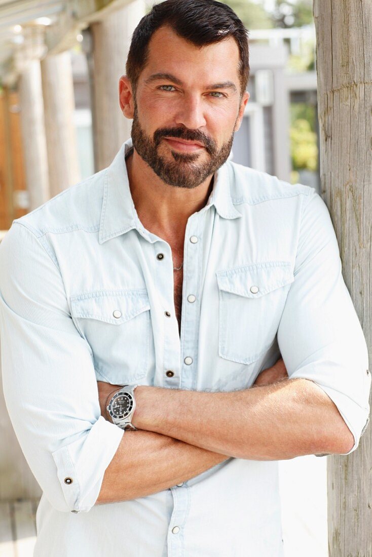 A dark-haired man with a beard wearing a light denim shirt on a terrace