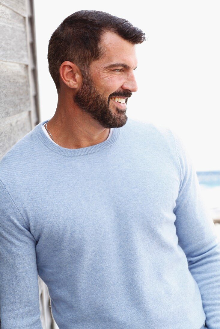 A dark-haired man with a beard wearing a light-blue jumper leaning against a wall