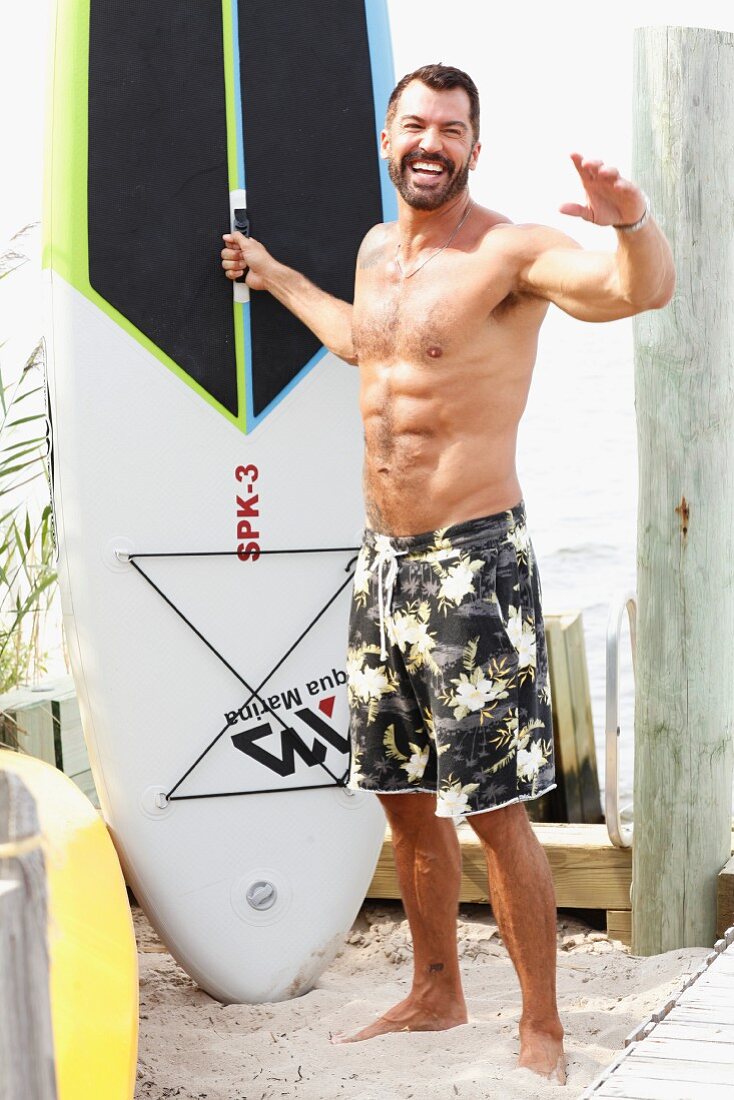 A dark-haired man on a beach wearing bathing shorts with a surfboard