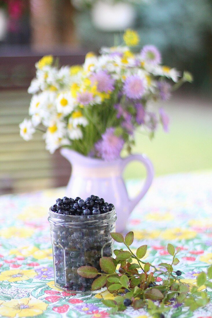 A jar of blueberries with a jug of meadow flowers on a garden table