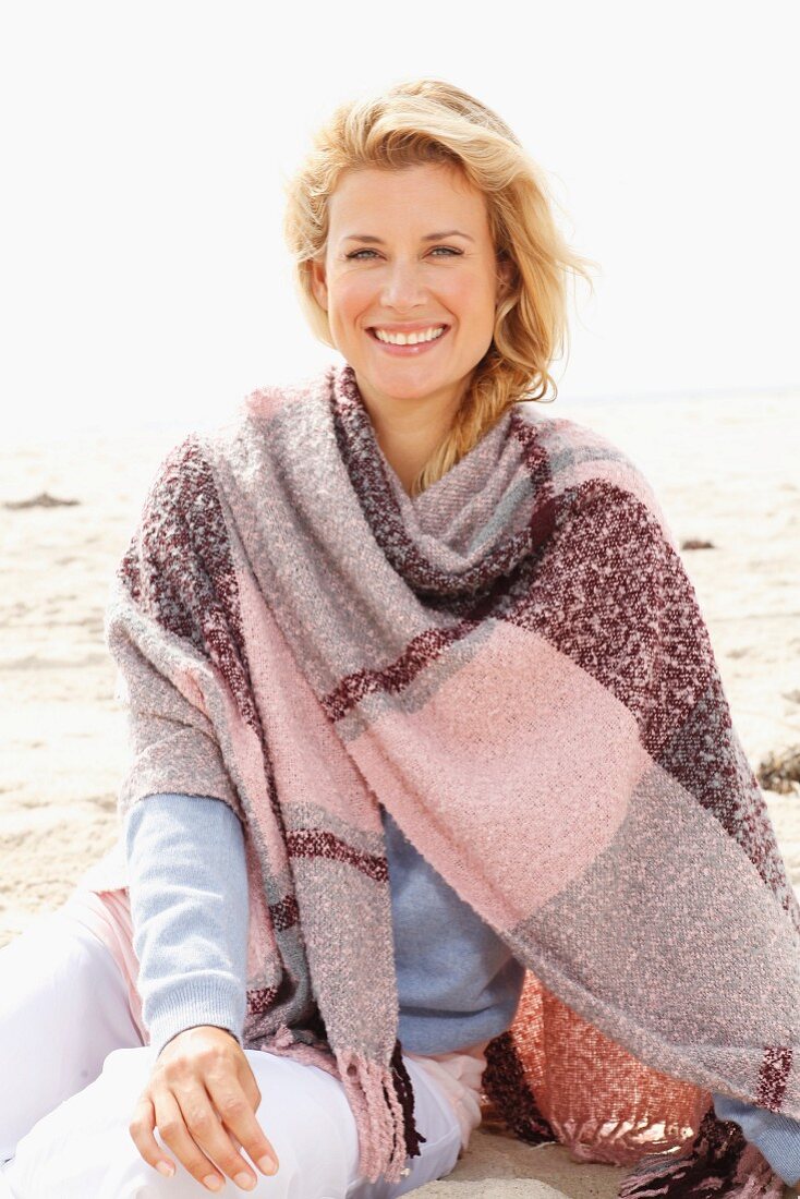 A blonde woman wearing a woollen shawl on a beach