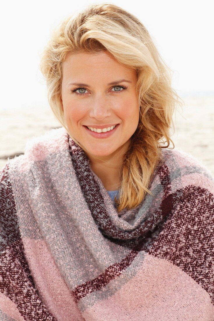 A blonde woman wearing a woollen shawl on a beach