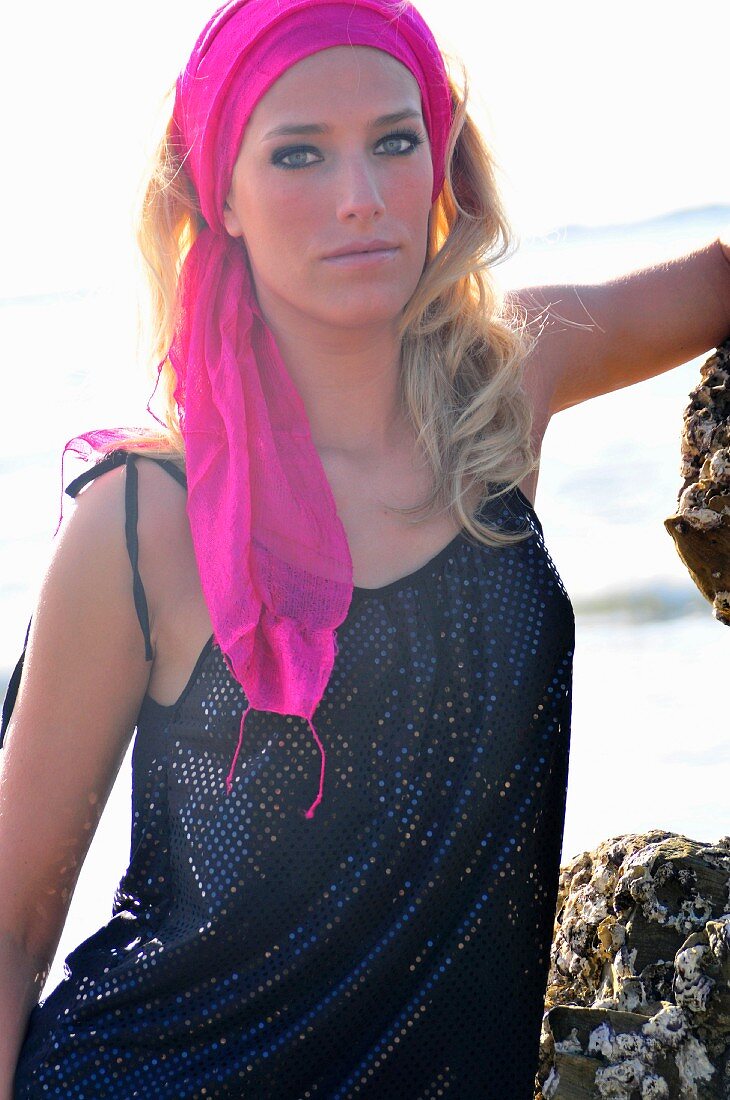 A blonde woman wearing a vest top standing on rocks by the sea