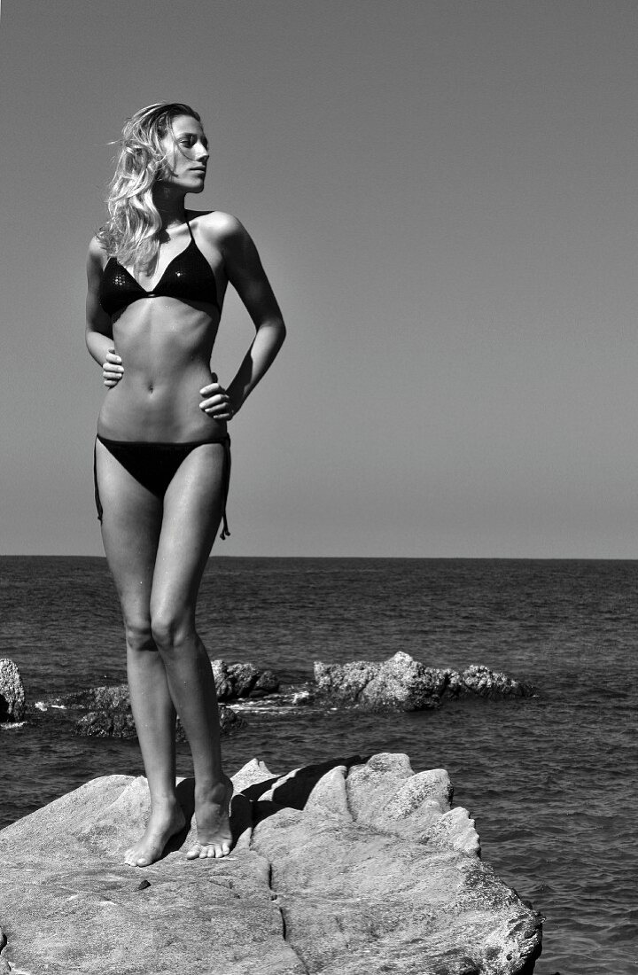 A blonde woman wearing a black sequinned bikini standing on rocks by the sea (black-and-white photo)
