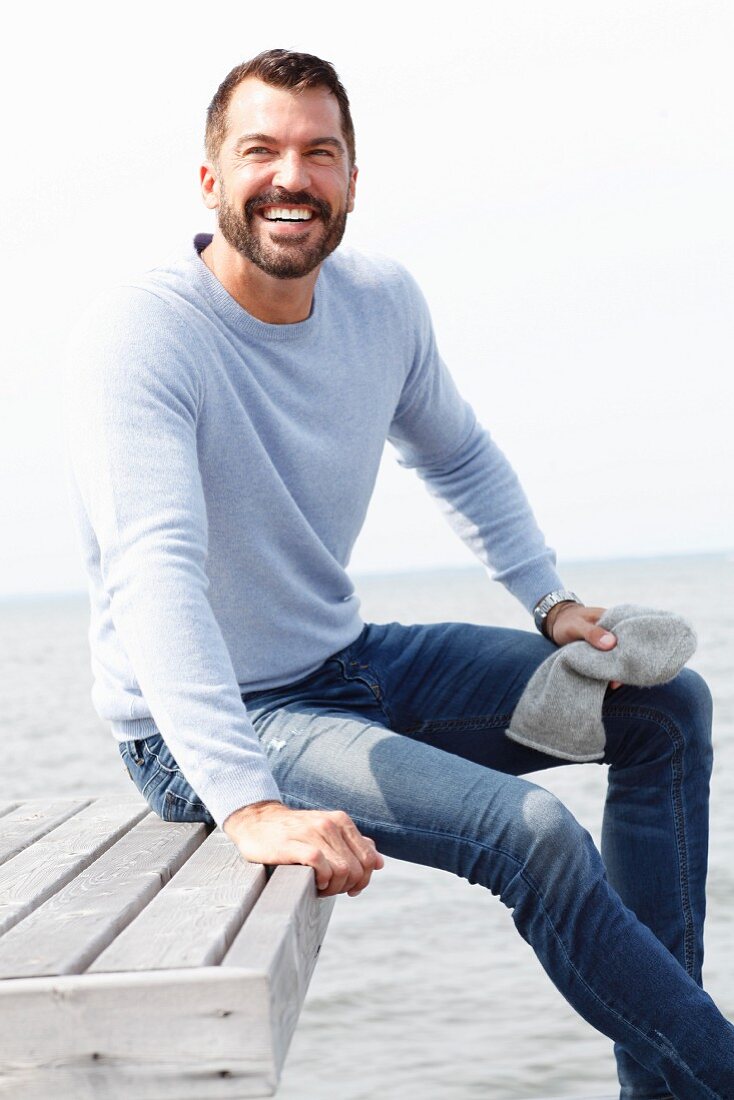 A man wearing a grey knitted hat, a light-blue jumper and jeans by the sea