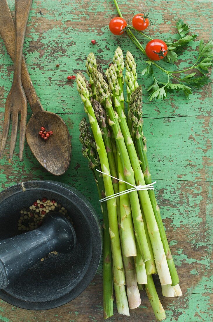 A bundle of fresh green asparagus, tomatoes and parsley on a wooden table, peppercorns in a mortar and salad servers