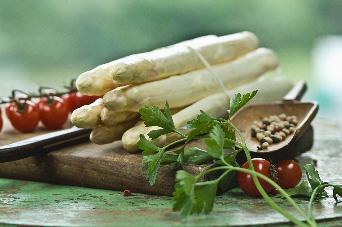 A bundle of white asparagus on a chopping board with a knife, peppercorns, parsley and cherry tomatoes