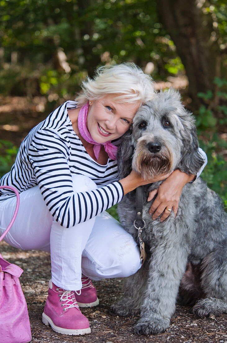 A blonde woman wearing a striped top, white trousers and pink shoes in a forest with a dog