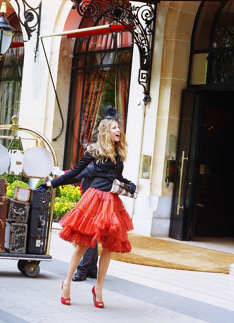 A blonde woman in front of a suitcase trolley wearing a red petticoat skirt, a black jacket and a fascinator