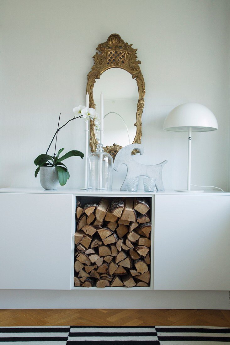 Sideboard mit glatten, weissen Fronten und Brennholzlager in Regalfach, Retroleuchte mit Halbkugelschirm und Antikspiegel an der Wand