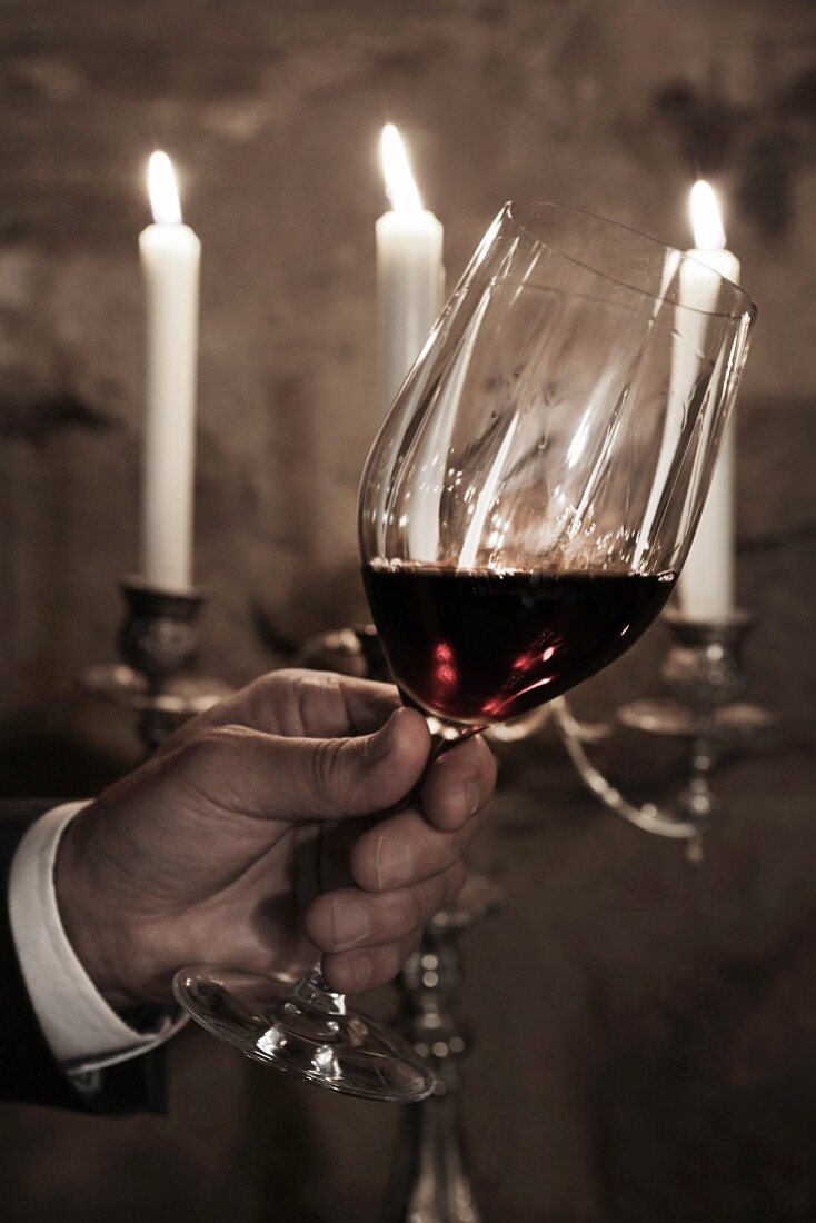 Weihnachten im Weinkeller: Hand hält Rotweinglas vor Kerzenleuchter