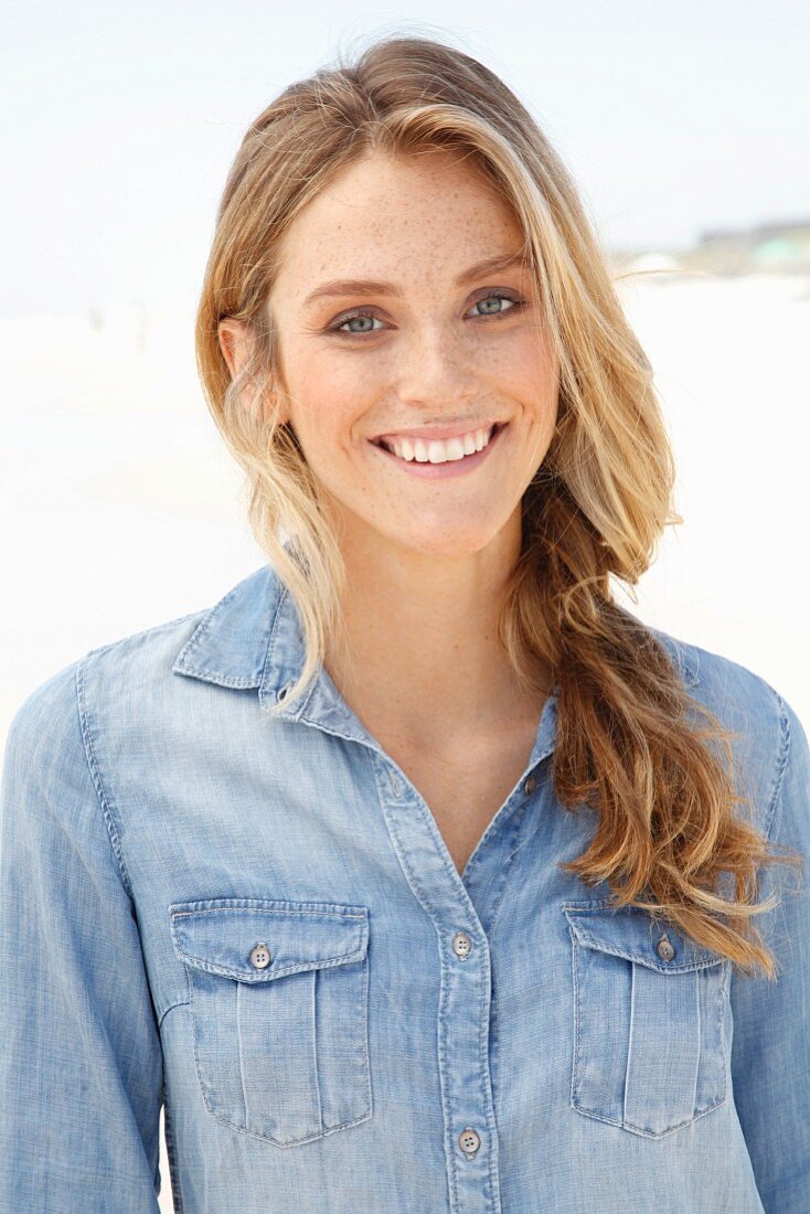 A young blonde woman on a beach wearing a denim shirt