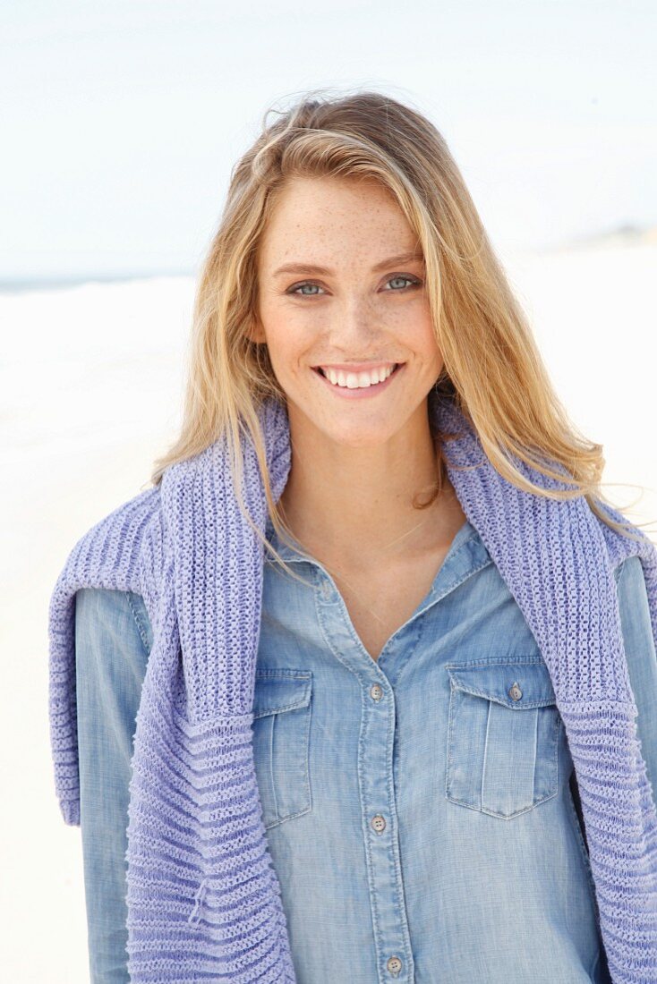 A young blonde woman on a beach wearing a denim shirt with a jumper over her shoulders