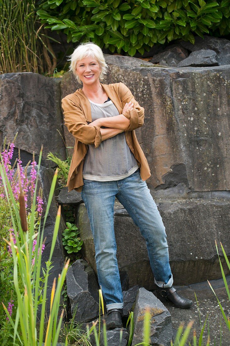 A blonde woman standing in a garden in front of rocks wearing casual clothing