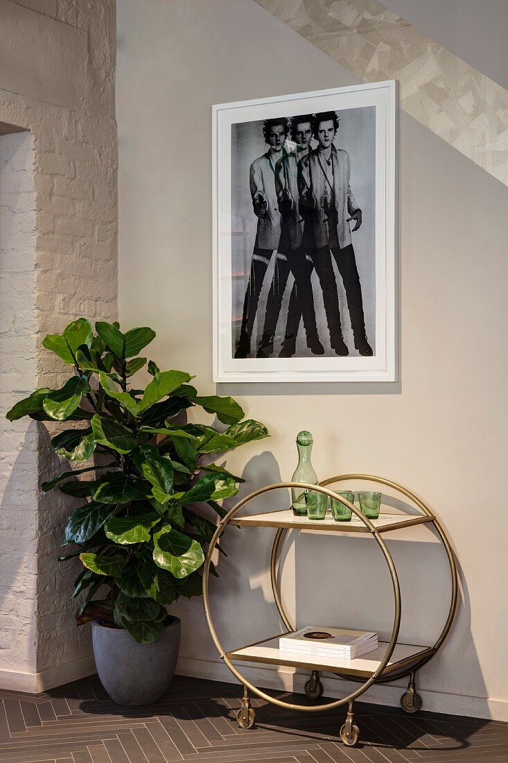 Green glasses and carafe on retro serving trolley, potted foliage plant and photo on wall
