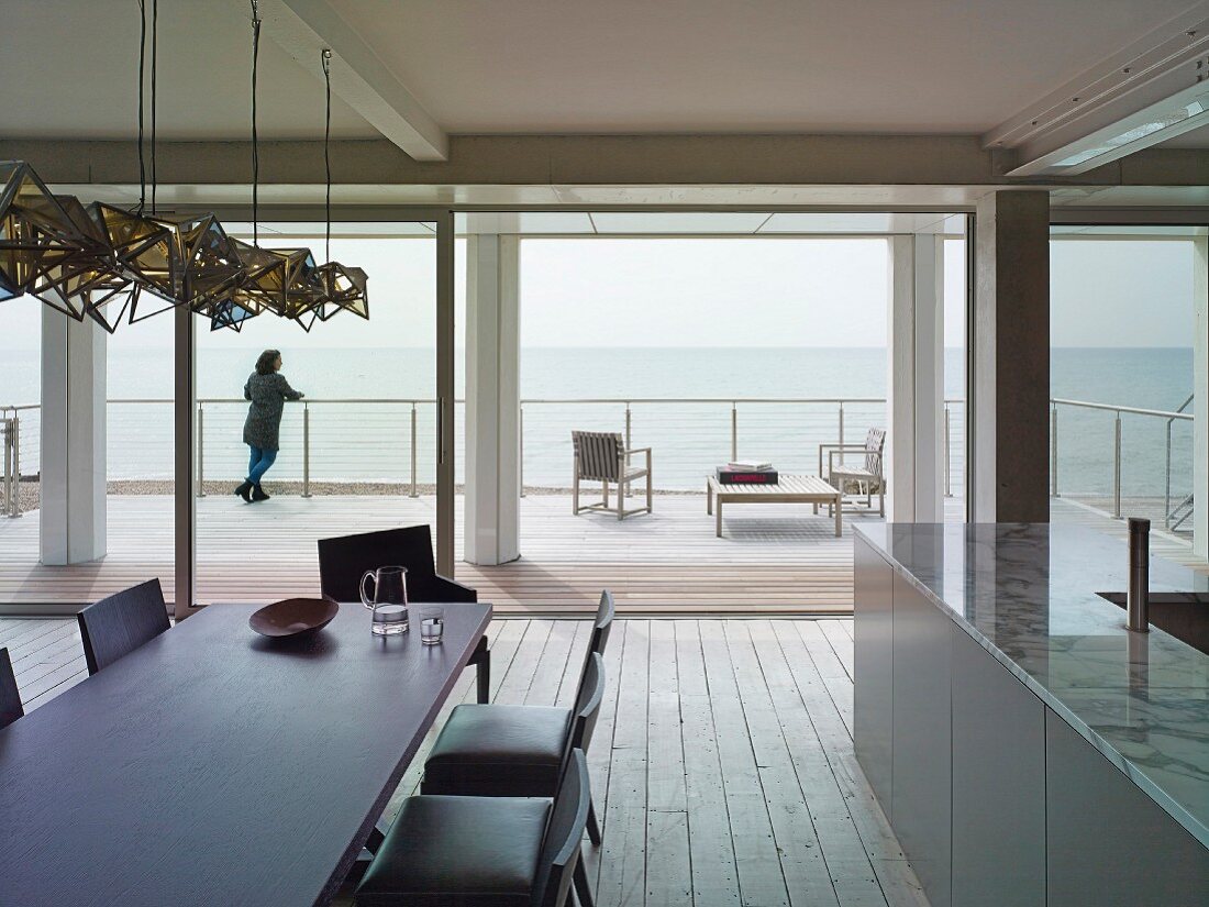 Blick vom Essplatz mit dunklem Tisch und Polsterstühlen durch offene Fensterschiebetür auf Holzterrasse und Meer