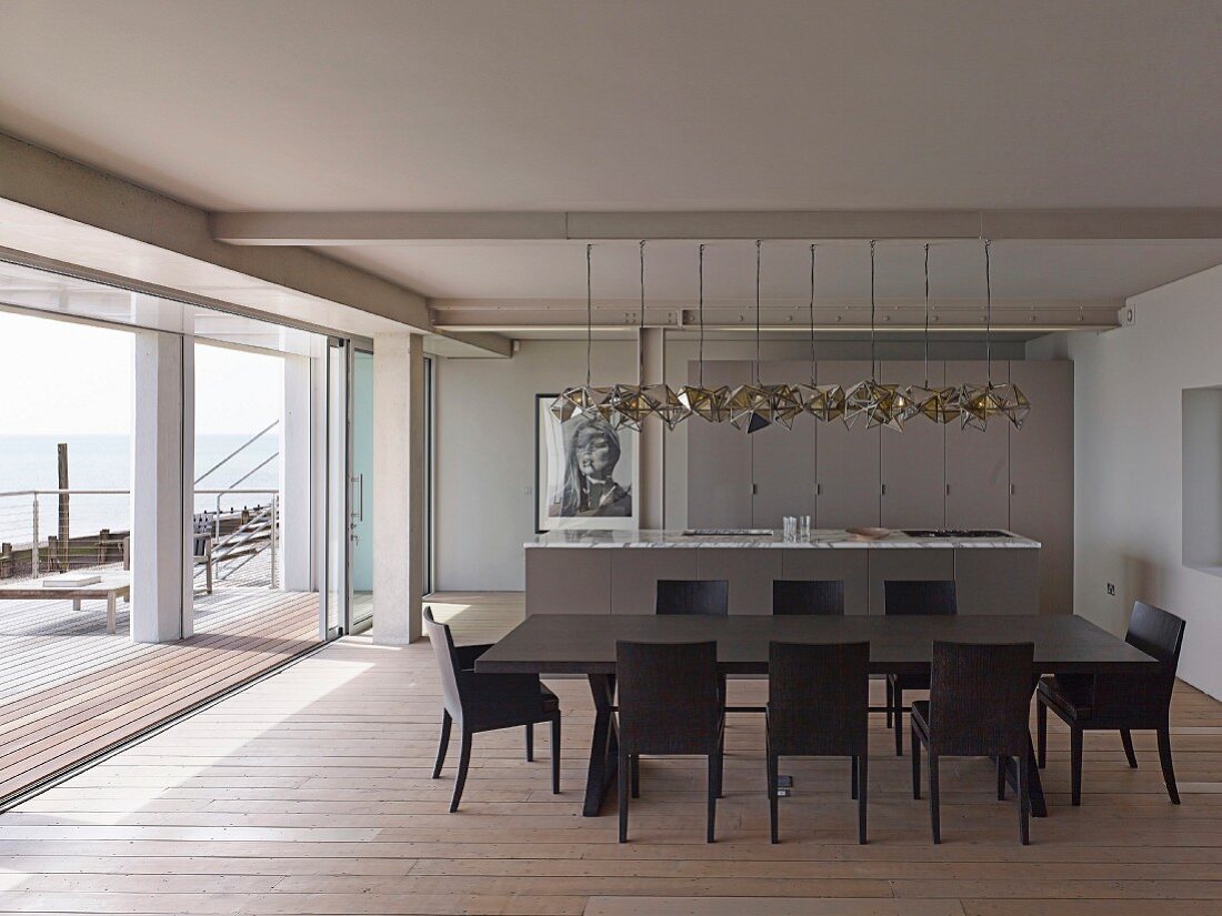 Dark dining table and matching chairs on wooden floor; kitchen counter and designer pendant lamps in open-plan interior with view of sea across terrace