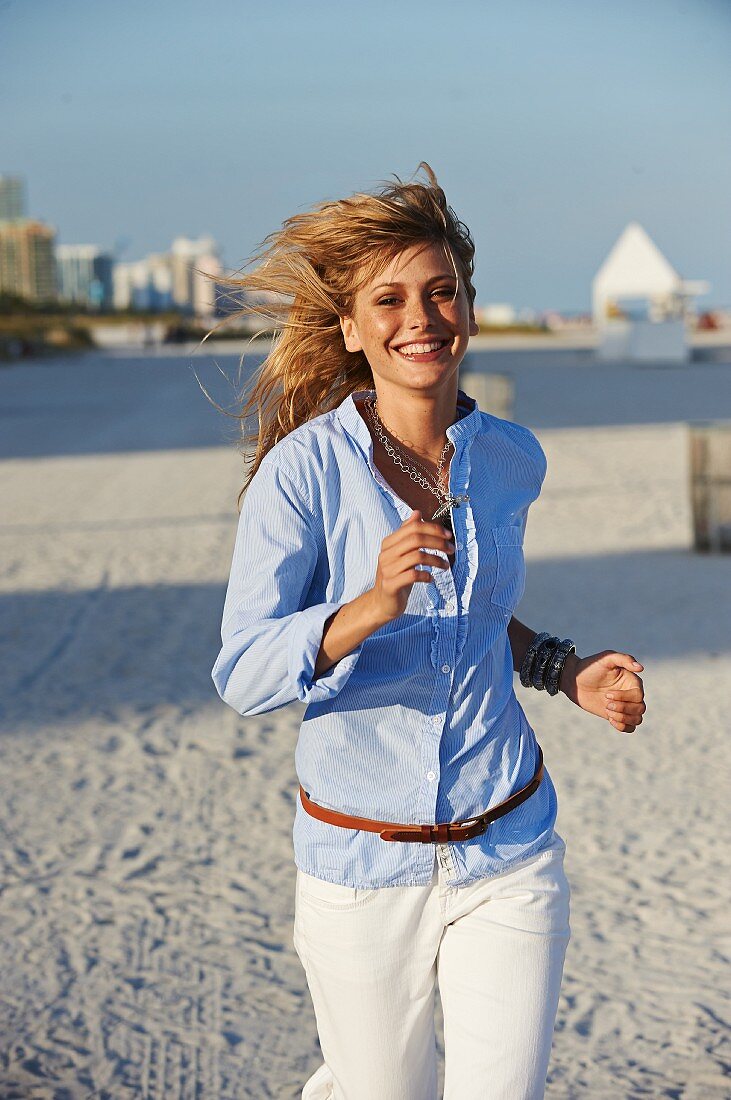 A young blonde woman jogging along a beach wearing a light blue shirt and white trousers