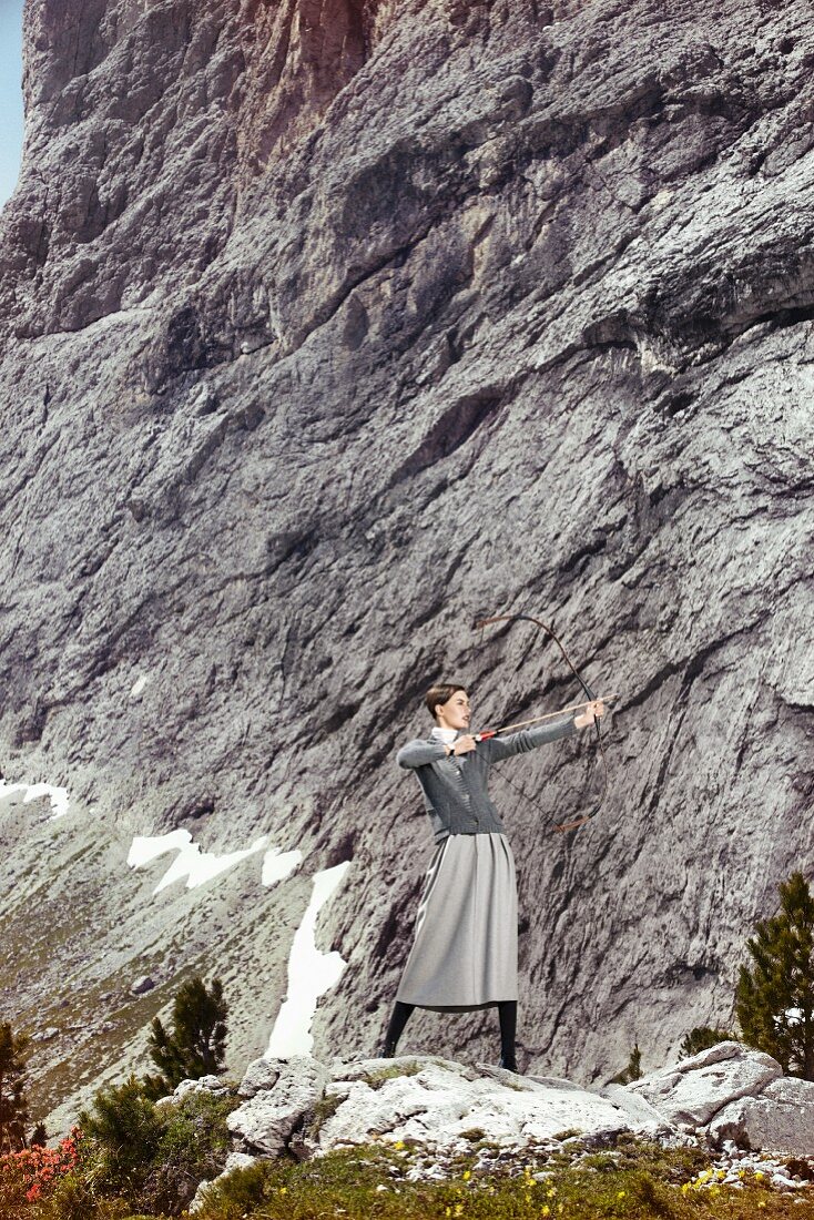 Kurzhaarige Frau in grauem Cardigan, Rollkragenpulli und knielangem Rock, mit Pfeil und Bogen