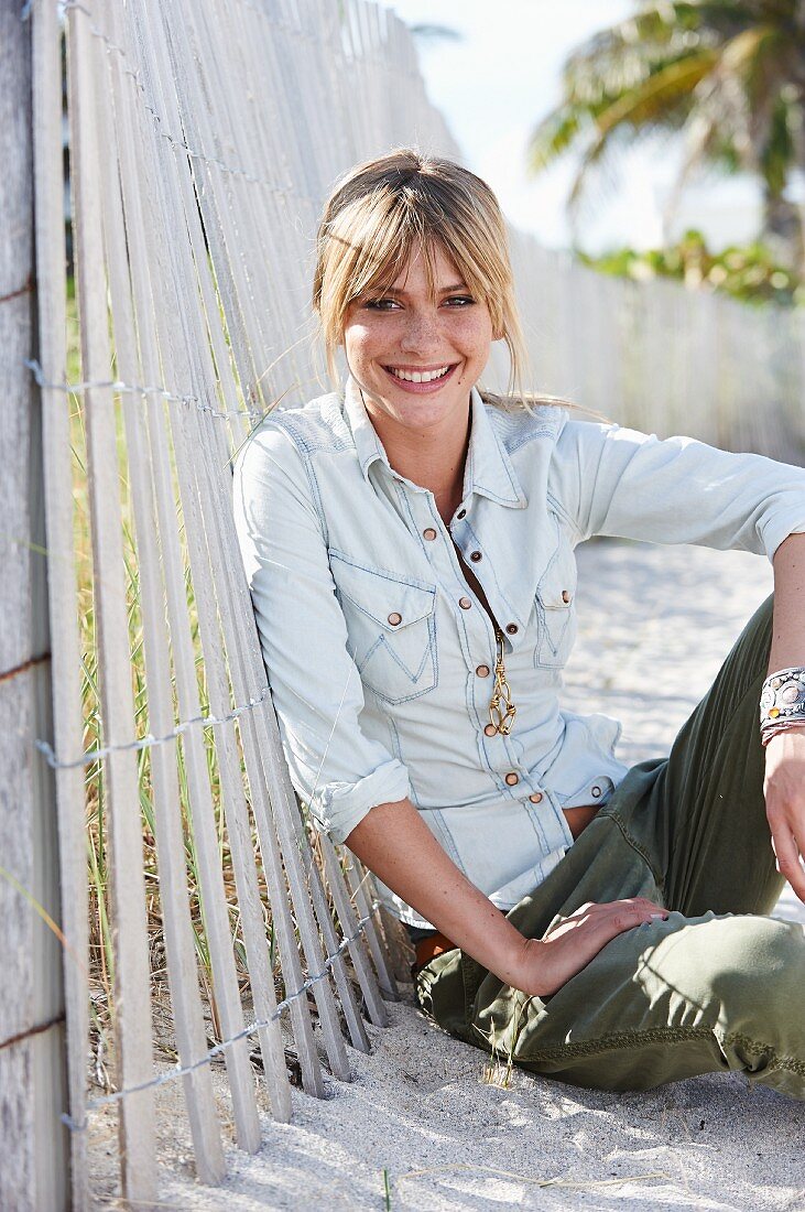 A young blonde woman sitting in the sand wearing a denim blouse and green trousers