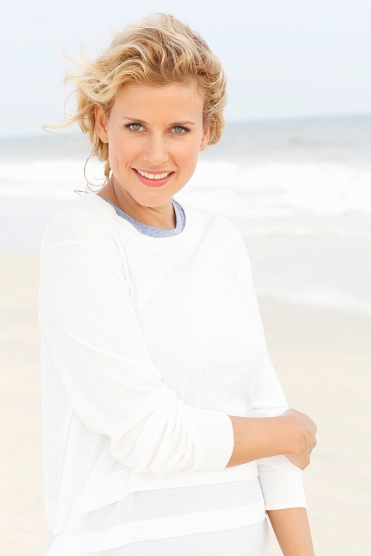 A blonde woman on a beach wearing a blue top and a white round-neck jumper