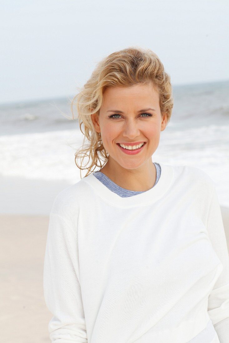 A blonde woman on a beach wearing a blue top and a white round-neck jumper