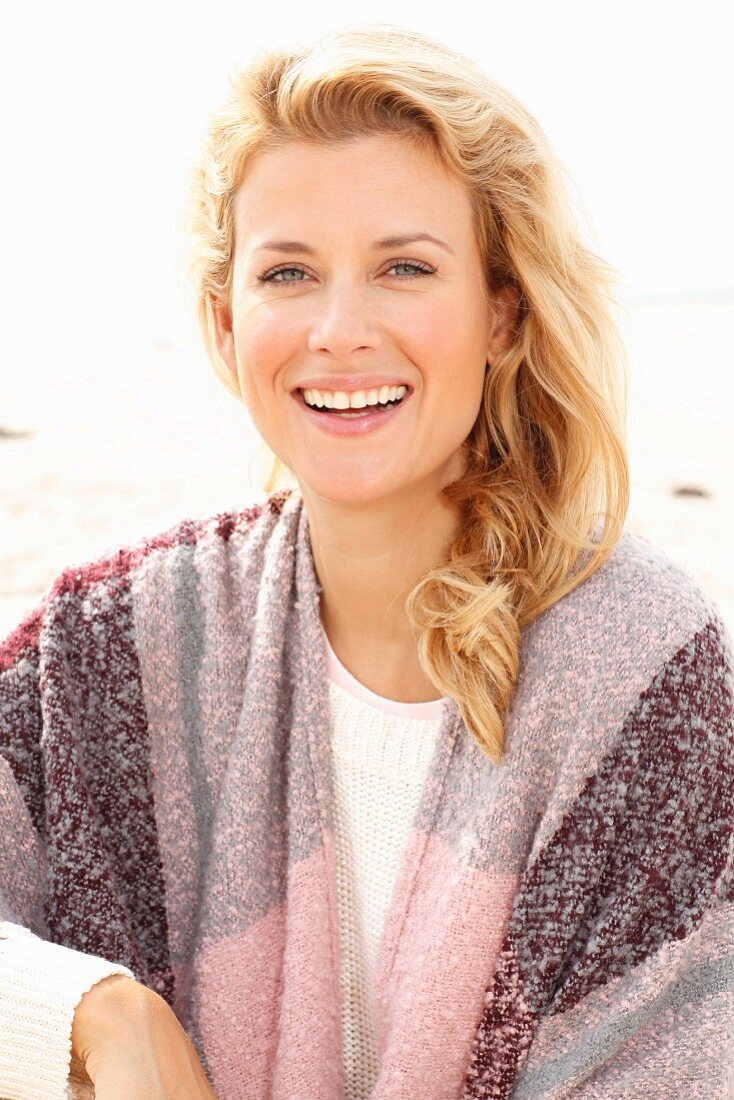 A blonde woman on a beach wearing a pink-toned shawl over her shoulders