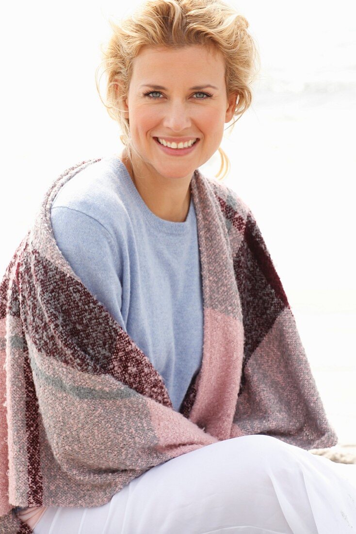 A blonde woman on a beach wearing a blue jumper and a pink-toned shawl over her shoulders