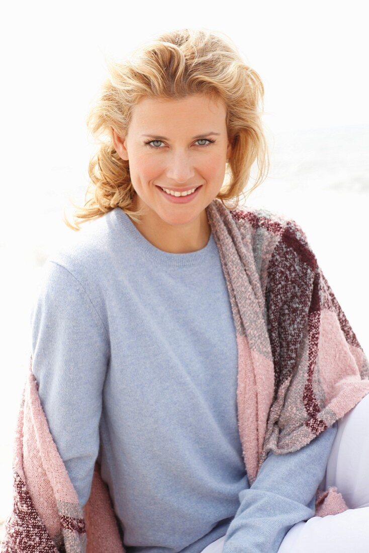 A blonde woman wearing a blue jumper and a pink-toned shawl over her shoulders