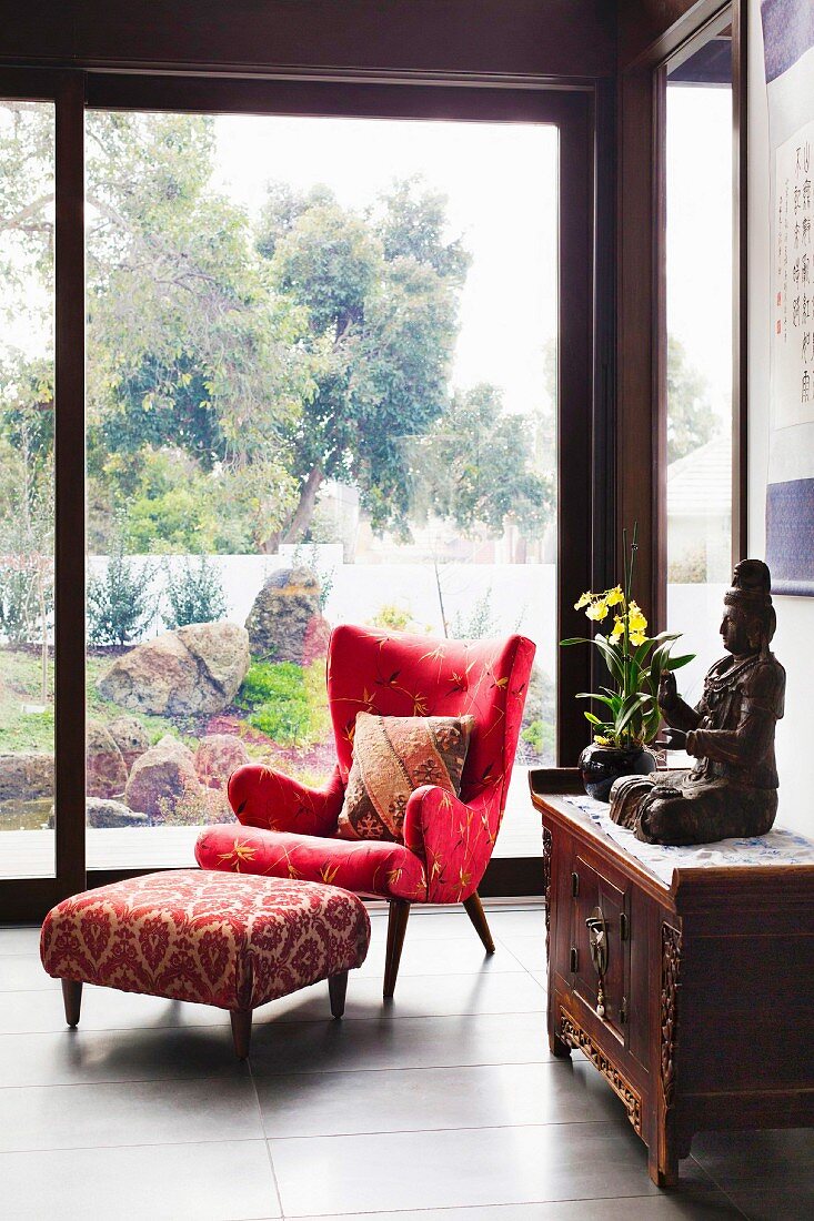 Elegant red reading armchair and footstool in front of floor-to-ceiling glazing with a garden view