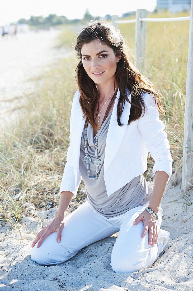 A brown-haired woman on a beach wearing a short, white jacket, a top and white trousers